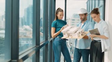 Proje yöneticisi ev modelini elinde tutarken, mimar mühendis takımı da ev tasarımı veya bina inşaatı hakkında fikir alışverişinde bulunuyor. Şehrin penceresinin yanında iç mimar duruyor. İzleme