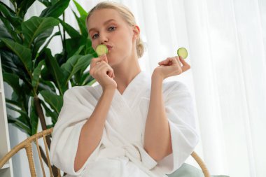 Elinde taze salatalık dilimleri tutan ve bornoz giyen bir kadın tatil köyünde ya da otelde lüks yüz derisi kaplıcalarının tadını çıkarıyor. Yüz ve güzellik tedavisi için cilt bakımı. Sessiz.