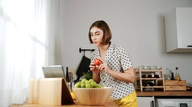 Kafkasyalı anne, modern mutfakta dizüstü bilgisayarda yemek tarifine bakarken salata yapmak için yemek hazırlıyor. Yetenekli ev bekçisi taze sebze kullanarak kahvaltı hazırlıyor. Pedagoji.