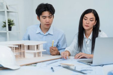 Profesyonel Asyalı erkek mimar ev modelinin uzunluğunu ölçmek için cetvel kullanırken, beyaz tenli genç meslektaşı ev modeliyle toplantı masasındaki verileri analiz etmek için dizüstü bilgisayar kullanıyor. Tertemiz..
