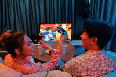 joystickli elleri çapraz oynayan bir çift arka tarafta TV ekranında dövüş oyunu oynuyor, modern rahat evinde neon ışıklı oturma odasında kanepesinde oturuyor. İnfobahn.