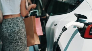 Yaz mevsiminde yeşil sürdürülebilir şehir bahçesinde ağır çekimde çalışan EV elektrikli araba yeşil sürdürülebilir şehirde yeniden şarj edilebilir elektrikli araç iç organları için alışveriş yapan genç çiftlerin yaşam tarzını gösteriyor.