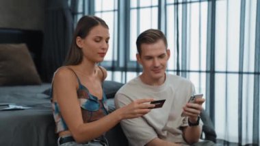 Genç çift, kredi kartıyla ödemek için internet üzerinden ödeme uygulaması ve dijital cüzdan kullanarak yatak odasında oturuyor. Mobil internet üzerinden alışveriş ve modern alımlar. Düzenle