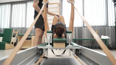 Eğitmen, müşteriye pilates reformatöründe rehberlik ediyor. Vücut geliştirme egzersizinde, vücut hareketlerine, esnekliğine ve beden kuvvetine odaklanmak için kayışlar kullanıyor. Asyalı kız pilates makinesinin önünde yatıyor. Alışkanlık.