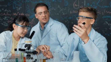 Yaratıcı öğretmen masadaki liseli çocukla mikroskop ve renkli sıvıyla test tüpüyle konuşurken kimyayı tahtaya doğrultuyor. BTMM sınıfında laboratuvar önlüğü giyen genç bir öğrenci. Öğretim.