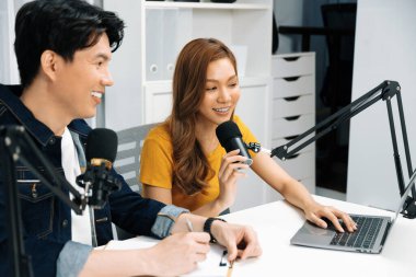 Smiling radio influencer hosts with channel social media online live streaming with talking topic collective question on script note and laptop to listeners at morning time at studio record. Infobahn. clipart