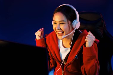 Yüzünde kulaklık renginde kulaklık takan, neon odada online oyun oynayan, Esport meziyetli takım oyuncularına yumruk kaldırarak oynayan güzel Asyalı kız flamasının sunucu kanalı. Taktik.