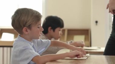 Beyaz tenli ilkokul öğrencisinin tahtada otururken bir nota bakması ve soruya cevap vermesi. Çocuklar derse katılırken genç öğretmen sınıf çalışmasını ya da sınavı açıklar. Pedagoji.