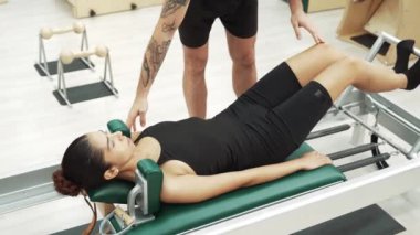 Kadın antrenör yardımıyla pilates egzersizi yapıyor. Pilates yaparken reformist makinesinde yatan ve çekirdek istikrarını arttırmak için ayak hareketlerinde yenilik yapan bir kız. Alışkanlık.