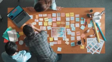 Gündelik kıyafet giyen, dizüstü bilgisayara bakan ve fikir paylaşan iş ekibinin zaman atlaması. Yapışkan notlar ve kulaklıkla masada çalışan ve renk paletini kontrol eden yöneticinin hava görüntüsü. Sempozyum.