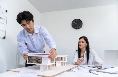 Professional male asian architect using ruler to measure house model length while young beautiful caucasian colleague using laptop to analyzed data on meeting table with house model. Immaculate. clipart