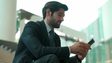 Gülümseyen yetenekli işadamı merdivenlerde otururken kameraya bakıyor. Genç profesyonel proje yöneticisi arka planda bulanık cep telefonunu tutarken kameraya gülümsüyor. Sevinçli..