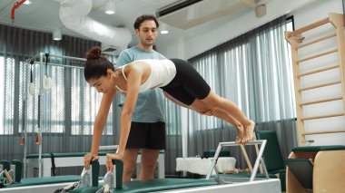 Bir pilates eğitmeni, müşteriye bir ıslah makinesinin tahta varyasyonunda rehberlik eder. Sağlıklı bir kadın, dengeyi geliştirmek için antrenör yardımı ile yenilikçi üzerinde yan kalas değişimi yapıyor. Alışkanlık.