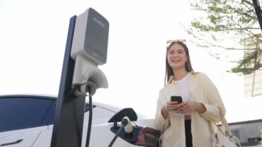 Ağır çekimde genç bir kadın, alışveriş merkezinin otoparkındaki şarj istasyonundan şarj olan EV 'nin pilini şarj ederken akıllı telefondan akü durumunu kontrol ediyor. Modern kadın elektrikli araçla alışverişe gider. Yararlı
