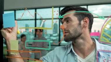 Akıllı proje yöneticisi cam duvara yapışkan notlar ve planlama stratejisi için zihin haritası yazarken, aynı zamanda çok kültürlü iş adamları da şehir manzaralı iş planlarını tartışıyorlar. İzleme