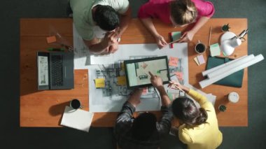İnşaat mühendislerinin masa başında proje planıyla otururken tablet kullanarak ev yapısını dizayn etmelerine tepeden aşağı bir bakış açısı. Ekip planlama ve inşaat hakkındaki fikirlerin havadan görüşü. Sempozyum.