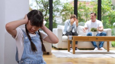 Stresli ve mutsuz genç kız köşeye kıvrılıp kulaklarını kapatarak arka planda ebeveyninin tartışmasını engelliyor. Evde aile içi şiddet ve travmatik çocukluk depresyona dönüşür. Eşzamanlama