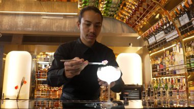 Asyalı barmen karışık kremalı votka bardağı servis ediyor. Önünde buzlu pembe çiçekli şeker, önünde özel içecek, lüks tezgah barında alkolü canlandırıyor. Vinosity.