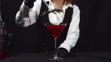 Makrografi, bir barmenin martini bardağına canlı kırmızı bir kokteyl karıştırma sanatına tanık olmak, güzel süslenmiş taze bir biberiye ve parlak siyah bir arka plan. Geliştirilebilir.