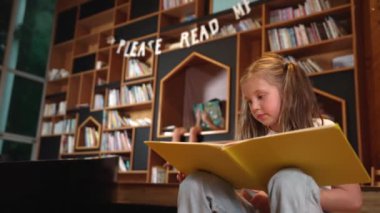 Genç, zeki, beyaz bir kız kütüphanede otururken kitap okuyor. Zeki çocuk öğrenme, okuma, kütüphanede kitap açma. Etkileyici çocuk geçmişi bulanık sayfayı çeviriyor. Etkinlik.