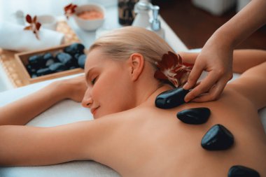 Lüks tatil köyündeki kaplıcalarda sıcak taş masajı. Gün ışığı sükunet ortamında, mutlu kadın müşteri bazalt taş masajından zevk alıyor. Rahatlatıcı bir sıcaklıkla vücudun üzerinde süzülüyor. Sessiz.