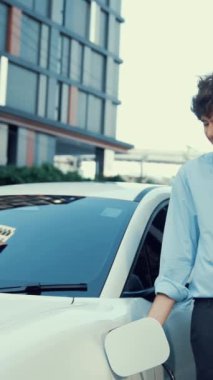 Takım elbiseli, hırslı bir işadamı elektrik kablosu ve yenilenebilir enerji enerjili bir elektrikli araçla şarj istasyonunda dururken elektrikli aracına yaslanıyor..