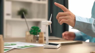 Temiz enerji ve ahşap blok kullanılarak temsil edilen yel değirmeni modelini izleyen iş adamı, gelecekteki yönetim için dizüstü bilgisayar ve defterle çalışan, eko ev dağınıklığını sembolize ediyor. Tanımlama.