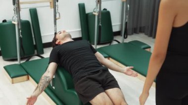 Erkek müşteri pilates ark fıçısında bacak kaldırma alıştırması yapıyor. Vücut dengesini, bacak dayanıklılığını ve spor salonundaki esnekliğini arttırmak için eğitmen yardımı ile birlikte. Öğrenci yalancı ve muziplik pozu veriyor. Alışkanlık.