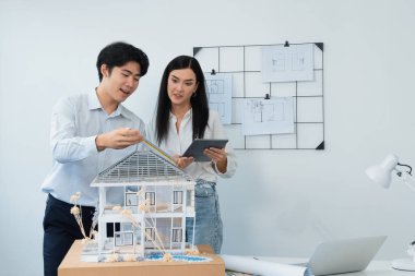 Güzel, genç, beyaz bir proje yöneticisi Asyalı mühendise, mühendislik masasında ev modelini ölçerken tablet kullanarak bina detaylarını anlattı. Tertemiz.