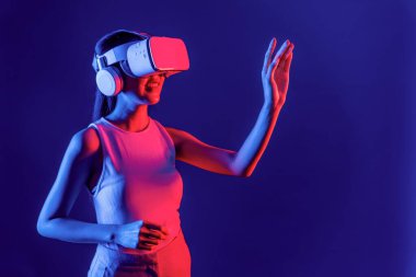 Çevresi siberpunk neon ışıklarıyla çevrili zeki bir kadın metaevrene bağlı VR kulaklık takıyor, gelecekçi siber uzay topluluğu teknolojisi. Sanal objeye el ile dokunan kadın. Halüsinasyon.