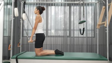 Kadın spor salonunda pilates reformatörüyle çekirdek güçlendirme egzersizi yapıyor. Çekici bayan pilates eğitmeni diz çökmüş vaziyette Cadillac 'ta omuz hareketliliğini arttırmak için bekliyor. Alışkanlık.