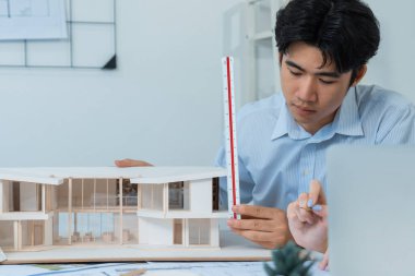 Professional male asian architect using ruler to measure house model length while young beautiful caucasian colleague using laptop to analyzed data on meeting table with house model. Immaculate. clipart