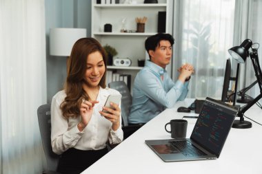 Profilde gülümseyen Asyalı güzel iş kadını, iş gününde modern ofiste bilgisayarla akıllı telefondan e-postaları kontrol ediyor. Bulanık arka plan çalışanı bilgisayardaki verileri analiz ediyor. İnfobahn.