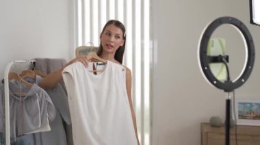 Genç sosyal medyum kadın moda videosu yapıyor. Blogcu kameraya selfie çubuğu ve ışık halkası kullanarak gülümserken, aynı zamanda ikna edici çevrimiçi kıyafetlerin izleyicilere veya takipçilere vlog satmasını sağlıyor. Düzenle
