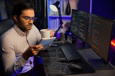 Akıllı bilişim geliştiricisi, bilgisayar ekranındaki yazılım geliştirme kodlaması üzerine kahveyle birlikte çalışıyor, en yeni uygulama projesi programı ile birlikte neon ışıklı siber ofiste çevrimiçi web sitesi verilerini güncelliyor. Tahmin et..