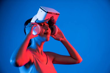 Zeki bir kadın gözünün üzerine VR bağlayan neon ışığı kulaklığı ile ayakta duruyor. Meta-evreni, gelecekteki siber uzay teknolojisini bağlayan cihaz. Elle gözlük tutan bir kadın. Halüsinasyon.