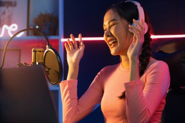 Genç ve güzel Asyalı yayıncının sunucu kanalı, dinleyicilerle sosyal medyada müzik dinliyor, pastel kulaklık takıyor, kapalı alanda neon ışıklı stüdyoda şarkı söylemek için mikrofon kullanıyor. Taktik.