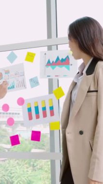 İş adamları ofisteki proje planlama kurulunda çalışıyor ve proje gelişimini analiz etmek için iş arkadaşlarıyla sohbet ediyorlar. Düzenli olması için cam duvara yapıştırılmış yapışkan notlar kullanıyorlar. .