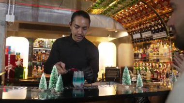 Lüks bar tezgahında kırmızı kurutulmuş meyve biberiyesiyle Noel mavisi lagün kokteylini süsleyen barmen ve VIP grup arkadaşı veya çift yeşil neon Noel ağacı festivaline ev sahipliği yapıyor. Vinosity.