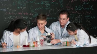 Çok kültürlü bir öğrenci mikroskop altında incelerken güzel bir kız da numuneyi inceliyor. BTMM sınıfında karatahta bilim teorisi deneyi yapan liseli çocuklara bakan öğretmen. Öğretim