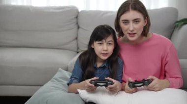 Mutlu anne ve enerjik kız oyun oynuyor ve birlikte vakit geçiriyorlar. Kafkasyalı anne, elinde joystick tutarken çocuğunun yanında oturuyor ve rahat oturma odasında kızıyla oyun oynamaya odaklanıyor. Pedagoji.
