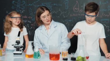 Öğretmen tavsiye verirken beyaz çocuk kimyasal sıvıyı karıştırıyor. Profesyonel eğitmen labaratuar kıyafeti giymiş, renkli solüsyonla dolu deney kabıyla sofrada çeşitli öğrencileri arıyor. Etkinlik.