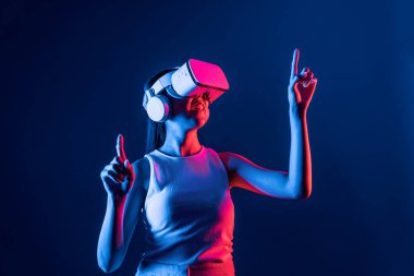 Siberpunk neon ışıklı cihazlarla çevrili zeki bir kadın metaevrene bağlı VR kulaklık takıyor, gelecekçi siber uzay teknolojisi. Sanal objeyi işaret eden bir kadın. Halüsinasyon.