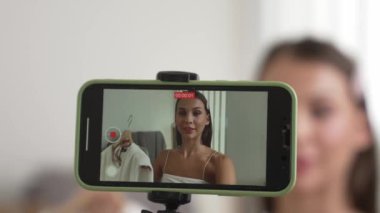 Genç sosyal medyum kadın moda videosu yapıyor. Blogcu kameraya selfie çubuğu ve ışık halkası kullanarak gülümserken, aynı zamanda ikna edici çevrimiçi kıyafetlerin izleyicilere veya takipçilere vlog satmasını sağlıyor. Düzenle