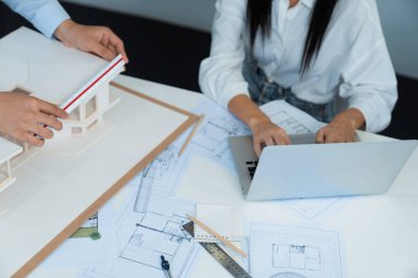 Profesyonel Asyalı erkek mimar ev modelinin uzunluğunu ölçmek için cetvel kullanırken, beyaz tenli genç meslektaşı ev modeliyle toplantı masasındaki verileri analiz etmek için dizüstü bilgisayar kullanıyor. Tertemiz..