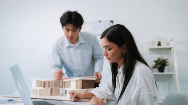 Profesyonel Asyalı erkek mimar ev modelinin uzunluğunu ölçmek için cetvel kullanırken, beyaz tenli genç meslektaşı ev modeliyle toplantı masasındaki verileri analiz etmek için dizüstü bilgisayar kullanıyor. Tertemiz..