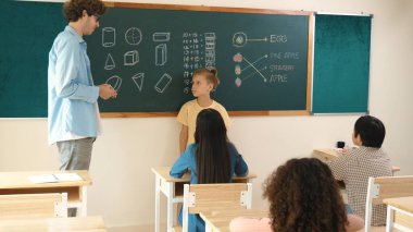 Profesyonel öğretmen konuşuyor ve sınıfta Asyalı çocuğa fikrini açıklıyor. Sınıfın önünde karatahta matematik dersiyle dikilmiş beyaz öğrenci dinleme eğitmeni. Pedagoji.