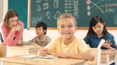 Sınıfta mutlu bir çok kültürlü öğrenci ilkokulda sınıfta sınav yaparken ya da not yazarken beyaz çocuk kameraya gülümsüyor. Eğitim konsepti. Pedagoji.