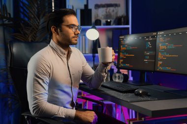 Akıllı IT geliştiricisi, bilgisayar ekranında yazılım geliştirme kodlu kahve fincanı tutuyor. Dışarıya bakıyor, program güncellemesi internet sitesi verilerini neon ışıklı siber ofiste sunuyor. Tahmin et..