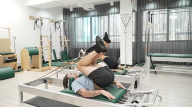 Müşteri, spor salonundaki yenilikçi bir makinede kayışla ters bacak kaldırma hareketi yapıyor. Çekirdek bağlantısını, dengesini ve esnekliğini geliştirmek için ters takla atan iki mutlu antrenör. Alışkanlık.